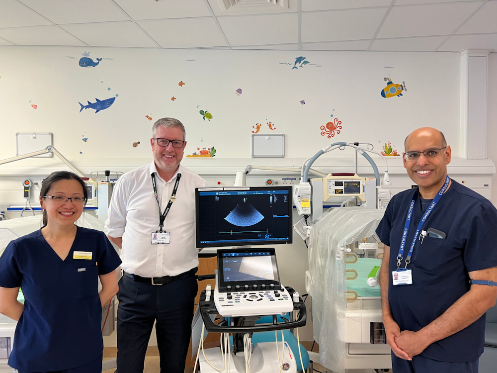 Three healthcare professionals in scrubs stand around an ultrasound machine in a hospital room. Their name badges are visible and starfish/ocean decals decorate the wall behind them, suggesting a pediatric or family-friendly setting.